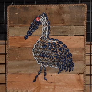 May include: A wooden wall art piece featuring a bird crafted from blue and silver bottle caps. The bird has a red bottle cap eye and is mounted on a wooden plank background. The artwork is framed with rope.