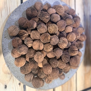 May include: A collection of brown walnuts fills a gray, circular dish. The walnuts have textured, ridged shells. The dish is set against a backdrop of light brown wooden planks, creating a rustic aesthetic.