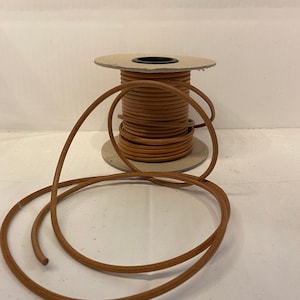May include: A spool of brown leather cord, wound around a cardboard spool. The cord is about 3/16 inch thick and is ready to be used for crafting projects.