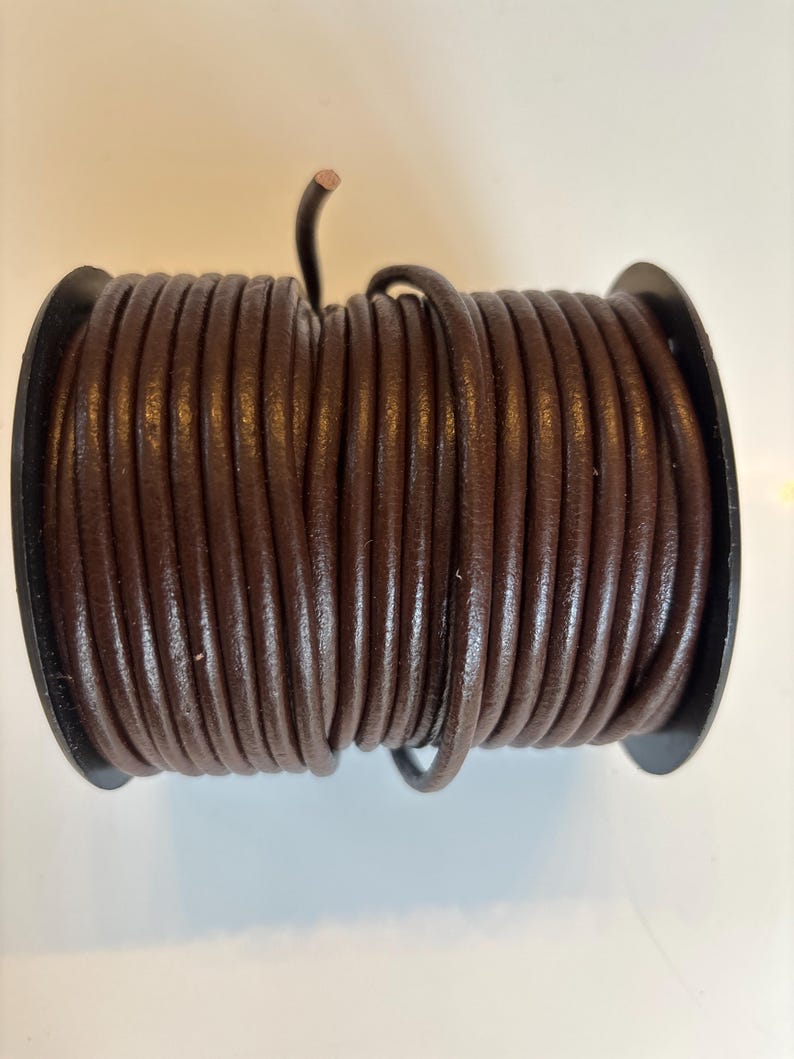 May include: A spool of brown leather cord. The cord is wound tightly around a black plastic spool.