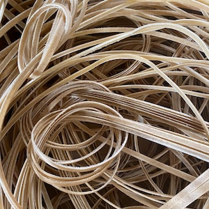 Op de afbeelding: Een verzameling dunne, lichtbruine fluwelen lintstroken gerangschikt in een verwarde, overlappende hoop. De linten hebben een zachte, pluizige textuur op het oppervlak en zijn in verschillende lussen gewikkeld, wat een dicht en gelaagd uiterlijk creëert.