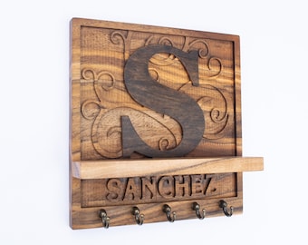 Può includere: Portachiavi in legno con una "S" marrone scuro e il nome "SANCHEZ" sotto un piccolo ripiano. Il portachiavi ha cinque ganci color bronzo e volute decorative intagliate nel legno.