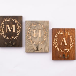 May include: Three wooden wall hooks with a leafy wreath design and a single initial in the center. The hooks are in dark brown, light brown, and reddish brown wood.