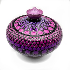 Puede incluir: Una caja redonda de madera con tapa, pintada con un diseño de mandala en tonos de rosa y morado. La tapa tiene un pequeño pomo puntiagudo.