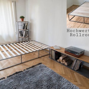 May include: A metal bed frame with a slatted base. The frame is made of black metal and has a simple, modern design. The bed frame is sitting on a wooden floor with a gray rug in front of it.