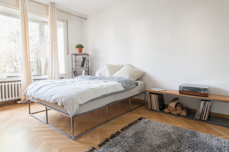 May include: A white bed with a metal frame sits in a bedroom with hardwood floors. The bed has a white duvet and two white pillows. A small wooden shelf with a record player and vinyl records sits in the corner of the room.