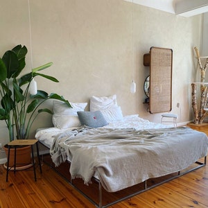 May include: A bedroom with a large bed with white linens and a gray throw blanket. There is a large green plant in a brown pot next to the bed. A small wooden stool is next to the plant. The room has light wood floors and a white wall with a woven wall hanging.