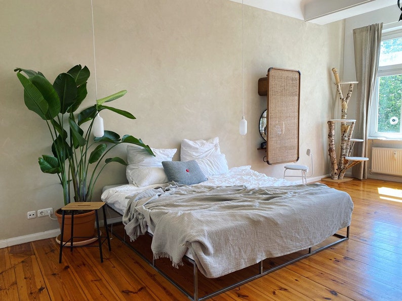May include: A bedroom with a large bed with white linens and a gray throw blanket. There is a large green plant in a brown pot next to the bed. A small wooden stool is next to the plant. The room has light wood floors and a white wall with a woven wall hanging.