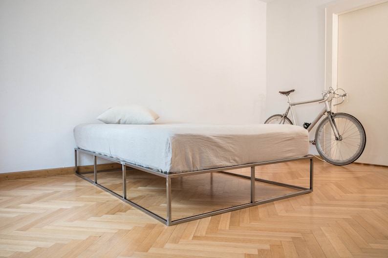May include: A white bed frame with a metal frame and a white mattress. The bed is in a room with a wooden floor and a white wall. A bicycle is leaning against the wall in the background.