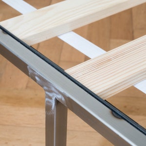 May include: Close-up of a metal bed frame with a slatted wooden base. The frame is silver and has a black rubber strip along the edge. The slats are made of light wood and are held together by white straps.