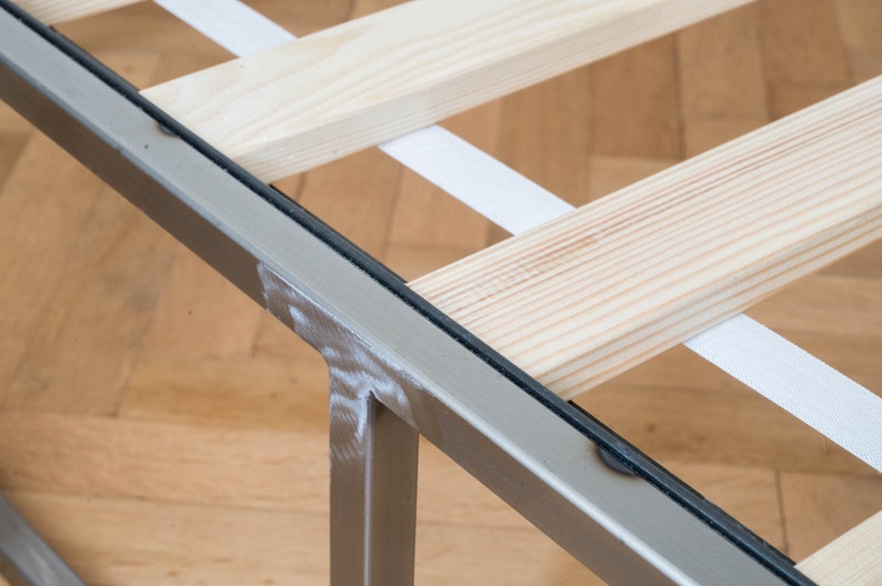 May include: Close-up of a metal bed frame with a slatted wooden base. The frame is silver and has a black rubber strip along the edge. The slats are made of light wood and are held together by white straps.