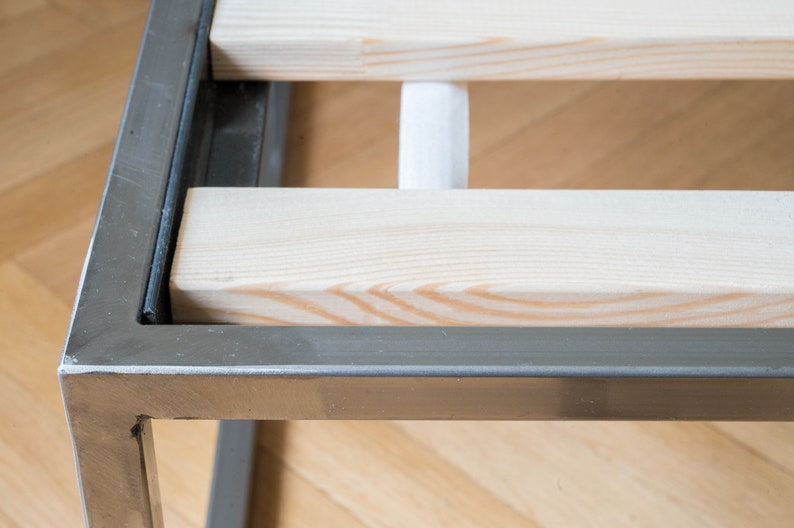 May include: Close-up of a metal bed frame with wooden slats. The frame is silver and the slats are light wood.