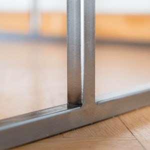 May include: Close-up of a metal table leg with a welded seam. The leg is made of silver-colored metal and has a rectangular shape.