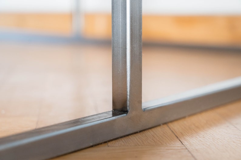 May include: Close-up of a metal table leg with a welded seam. The leg is made of silver-colored metal and has a rectangular shape.