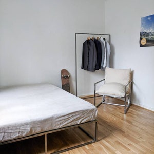 May include: A white bed frame with a white linen mattress, a black skateboard leaning against the frame, a black metal clothing rack with several jackets hanging on it, a white armchair with a white cushion, and a poster on the wall.