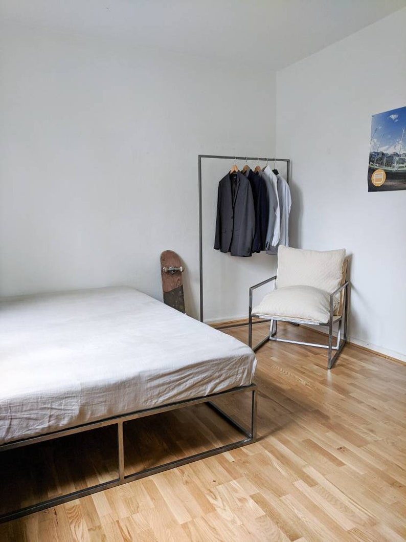 May include: A white bed frame with a white linen mattress, a black skateboard leaning against the frame, a black metal clothing rack with several jackets hanging on it, a white armchair with a white cushion, and a poster on the wall.