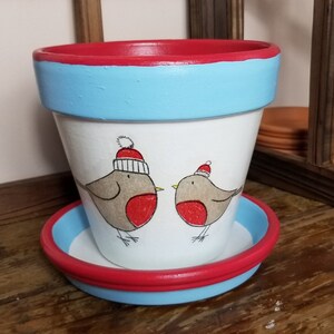 Peut inclure: Un pot en céramique blanche avec un bord rouge et bleu, orné d'un motif peint à la main de deux rouges-gorges portant des bonnets de Père Noël. Le pot a une soucoupe assortie.