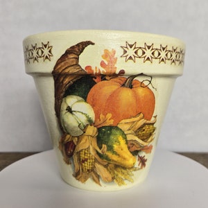 May include: A cream-colored ceramic planter with a fall harvest design. The planter features a cornucopia overflowing with pumpkins, gourds, and corn. The rim of the planter is decorated with a brown and white star pattern.