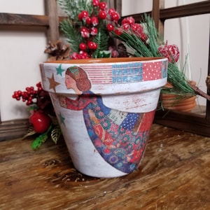 May include: A white ceramic planter with a distressed finish, featuring a colorful angel design with stars and patchwork patterns. The planter is decorated with red, blue, and white colors.