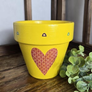 May include: A bright yellow flower pot with a red heart design. The heart has a patterned texture. The pot has small, colorful accents and is sitting on a wooden surface. A green plant is next to the pot.