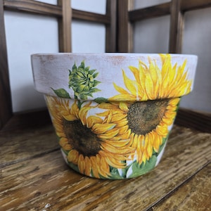 May include: A white ceramic plant pot adorned with vibrant yellow sunflowers and green foliage. The pot has a distressed, aged look. The sunflowers are in full bloom, with detailed petals and dark brown centres. A green sunflower bud is also visible.