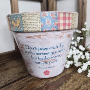 May include: A white painted flower pot with a decorative rim featuring a butterfly, floral, and gingham pattern. The pot has a quote: "Don't judge each day by the harvest you reap, but by the seeds that you plant."