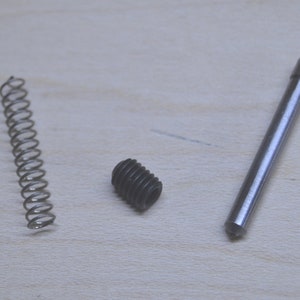 May include: A close-up of three metal parts: a spring, a small screw, and a long, thin rod with a pointed tip.