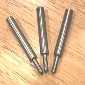 May include: Three silver metal awls with pointed tips. The awls are arranged in a V-shape on a wooden surface.