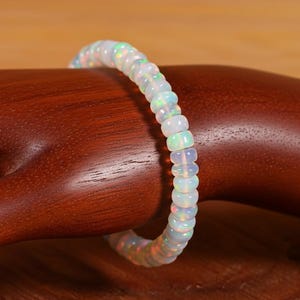 May include: A white opal bracelet with a beaded design. The bracelet features small, round opal beads, displaying a play of colour. The bracelet is shown on a wooden surface.