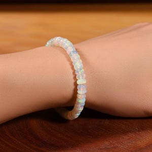 Peut inclure: Un bracelet en opale composé de petites perles rondes et iridescentes. Le bracelet est porté sur un poignet, mettant en valeur les couleurs subtiles des opales. Les perles semblent être enfilées, créant un bijou souple et élégant. Circonférence: environ 17 cm.