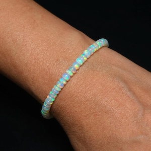 May include: A bracelet composed of small, round, translucent opal beads. Each bead displays a milky white base color with iridescent flashes of green, blue, and orange light. The beads are strung together in a single continuous line around the wrist.