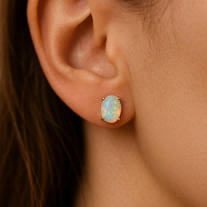 Könnte beinhalten: Ovale Opal-Ohrstecker mit Goldfassung. Die Ohrringe sind vor dem Hintergrund eines Ohrs und der Haut einer Person platziert. Der Opal hat eine milchig-weiße Basis mit Farbflecken.