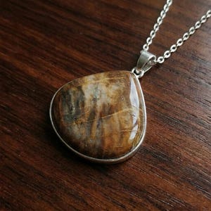 Puede incluir: Un colgante en forma de lágrima con una piedra pulida en tonos marrones y crema con vetas naturales visibles. La piedra está engastada en un bisel de metal plateado y cuelga de una delicada cadena de metal plateado.