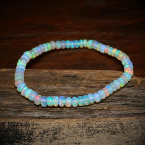 Pode incluir: Uma delicada pulseira composta por pequenas contas redondas de opala iridescente. As contas exibem uma gama de cores, incluindo branco, azul, rosa e verde, criando um efeito brilhante. A pulseira é exibida em uma superfície de madeira rústica.