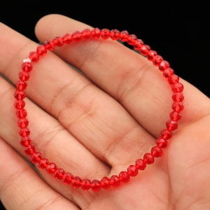 May include: A delicate red beaded bracelet. The bracelet is composed of small, faceted beads strung together, creating a continuous loop. The beads are a vibrant red color, and the bracelet is shown resting on a hand.