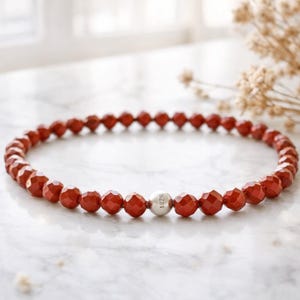 May include: A red beaded bracelet with faceted beads. The bracelet has a single silver bead with the text "1926" on it. The bracelet is displayed on a white marble surface.