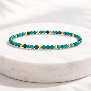 May include: A turquoise beaded bracelet with gold-colored accents. The bracelet is made of small, round beads and is displayed on a white marble surface. The beads are a mix of turquoise and dark green.