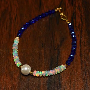 May include: A delicate bracelet featuring a combination of blue faceted beads, iridescent opal beads, and a central white pearl. The bracelet is accented with gold-colored metal and has a clasp closure. The jewelry is displayed on a dark wooden surface.