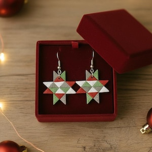 May include: A pair of star-shaped earrings with red, white, and green geometric patterns, displayed in a red velvet box. The earrings have silver hooks. The background includes Christmas ornaments and string lights, suggesting a holiday theme.
