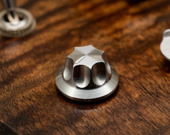 May include: Close-up of a silver metal knob with a star-shaped top. The knob is on a dark brown wooden surface.