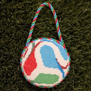 May include: A round, padded ornament featuring a cross-stitched design with abstract red, blue, and green shapes on a white background. The ornament is edged with a twisted rope trim in red, blue, and green, and includes a matching rope hanging loop.