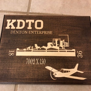 K&ouml;nnte beinhalten: Ein dunkelbraunes Holzschild mit einer wei&szlig;en Silhouette einer Flughafenlandebahn und einem kleinen Flugzeug. Das Schild tr&auml;gt die Aufschrift "KDTO DENTON ENTERPRISE" und "7002 X 150".