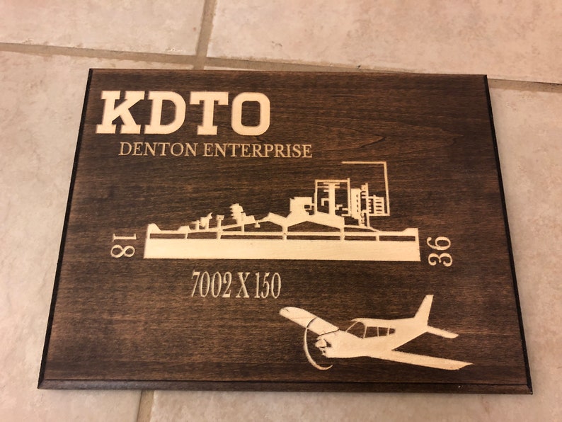 K&ouml;nnte beinhalten: Ein dunkelbraunes Holzschild mit einer wei&szlig;en Silhouette einer Flughafenlandebahn und einem kleinen Flugzeug. Das Schild tr&auml;gt die Aufschrift "KDTO DENTON ENTERPRISE" und "7002 X 150".