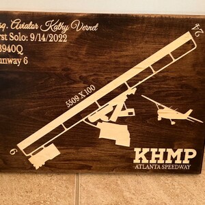 K&ouml;nnte beinhalten: Ein Holzschild mit braunem Hintergrund und wei&szlig;em Text. Das Schild zeigt eine Silhouette eines Flugzeugs und einer Landebahn. Der Text auf dem Schild lautet "Esq. Aviator Kathy Vernet", "First Solo: 9/14/2022", "N8940Q", "Runway 6", "5509 X 100", "KHMP" und "ATLANTA SPEEDWAY".