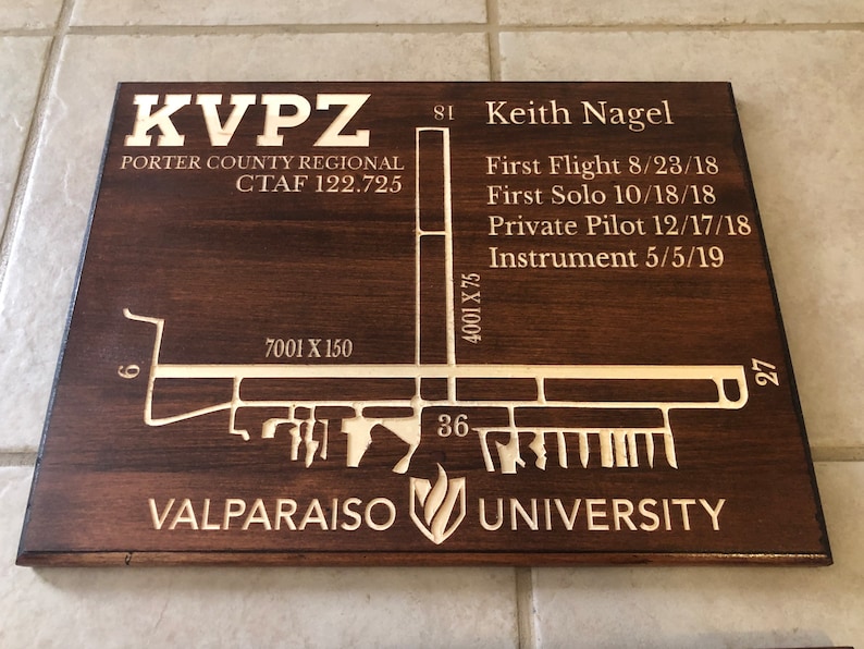 K&ouml;nnte beinhalten: Eine Holzplakette mit brauner Beize und einem hellbraunen, gravierten Design. Die Plakette zeigt ein Flugplatzlayout mit dem Text "KVPZ Porter County Regional CTAF 122.725" und "Valparaiso University". Die Plakette enth&auml;lt auch den Text "Keith Nagel" und Flugmeilensteine.