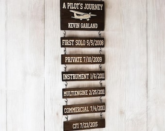 May include: A wooden sign with the text "A Pilot's Journey" and a small airplane graphic. The sign is hanging from a wall and has several smaller signs hanging below it with dates and text such as "First Solo 5/5/2008", "Private 7/10/2009", "Instrument 1/6/2011", "Multiengine 2/25/2011", "Commercial 7/4/2011", and "CFI 7/23/2015".