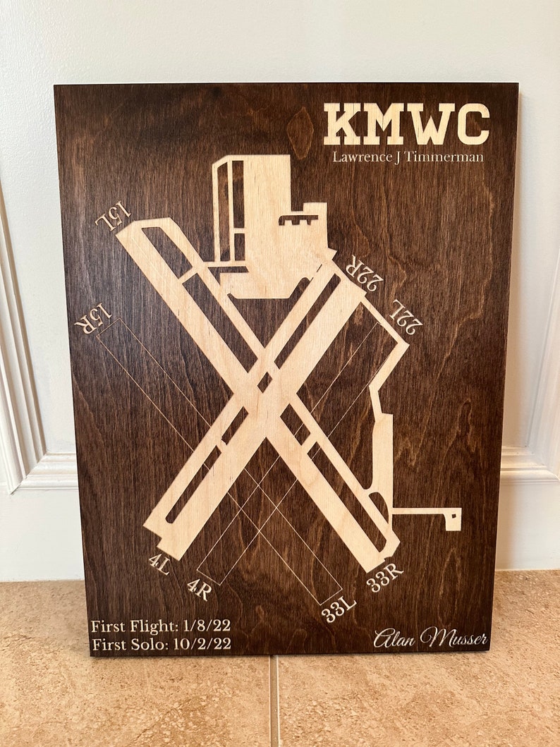 K&ouml;nnte beinhalten: Eine Holzplakette mit braunem Hintergrund und einem hellbraunen Design einer Flughafenlandebahn. Die Plakette enth&auml;lt den Text "KMWC Lawrence J Timmerman 22R 767 33L 33R 4L 4R First Flight: 1/8/22 First Solo: 10/2/22 Alan Musser".