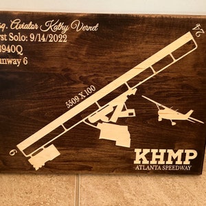 K&ouml;nnte beinhalten: Ein braunes Holzschild mit einer wei&szlig;en Silhouette eines Flugzeugs und einer Landebahn. Das Schild tr&auml;gt die Aufschrift "KHMP Atlanta Speedway" und den Text "Esq. Aviator Kathy Vernet First Solo: 9/14/2022 N3940Q Runway 6 5509 X 100 24".