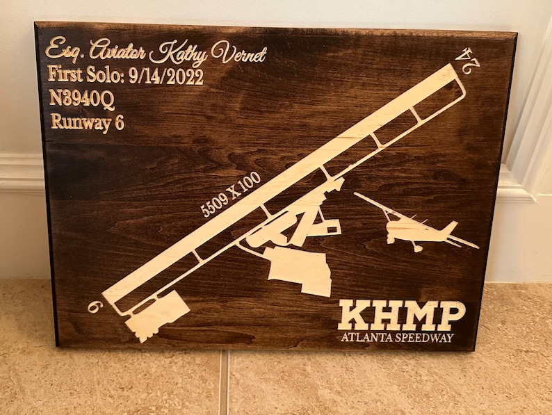 K&ouml;nnte beinhalten: Ein braunes Holzschild mit einer wei&szlig;en Silhouette eines Flugzeugs und einer Landebahn. Das Schild tr&auml;gt die Aufschrift "KHMP Atlanta Speedway" und den Text "Esq. Aviator Kathy Vernet First Solo: 9/14/2022 N3940Q Runway 6 5509 X 100 24".