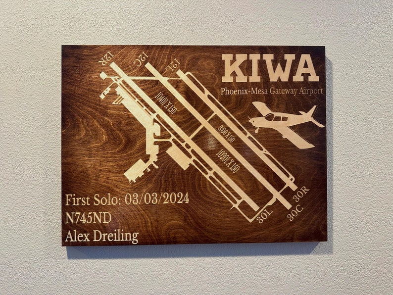 K&ouml;nnte beinhalten: Eine Holzplakette mit braunem Hintergrund und einem hellbraunen Flughafen-Start- und Landebahndesign. Die Plakette tr&auml;gt den Text "KIWA Phoenix-Mesa Gateway Airport" und "First Solo: 03/03/2024 N745ND Alex Dreiling".
